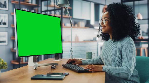 istock Latina Female Specialist Working on Desktop Computer with Green Screen Mock Up Display at Home Living Room while Sitting at a Table. Freelancer Female Chatting Over the Internet on Social Networks. 1313422650