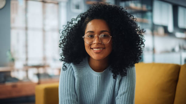 Portrait of a Beautiful Authentic Latina Female with Afro Hair Wearing Light Blue Jumper and Glasses. She Looks to the Camera and Smiling Charmingly. Successful Woman Resting in Bright Living Room. Portrait of a Beautiful Authentic Latina Female with Afro Hair Wearing Light Blue Jumper and Glasses. She Looks to the Camera and Smiling Charmingly. Successful Woman Resting in Bright Living Room. non binary gender photos stock pictures, royalty-free photos & images