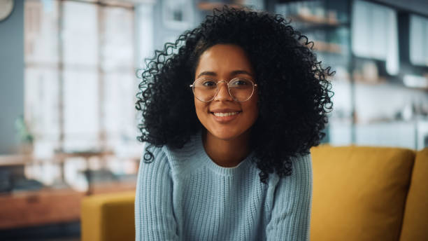 retrato de uma linda mulher latina autêntica com cabelo afro vestindo macacão azul claro e óculos. ela olha para a câmera e sorri encantadoramente. mulher bem sucedida descansando na sala de estar brilhante. - adult adults only assistant occupation - fotografias e filmes do acervo