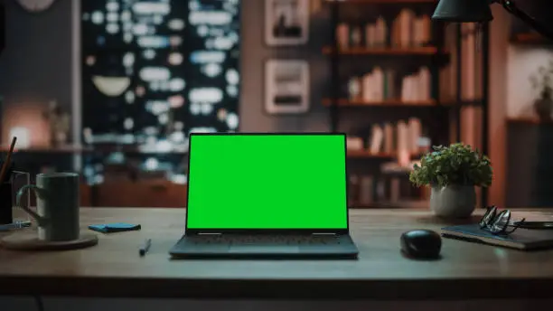 Laptop Computer with Mock Up Green Screen Display Standing on the Table in Cozy Living Room. In the Background Stylish Modern Home Office Studio in the Evening with Big Cityscape Window.