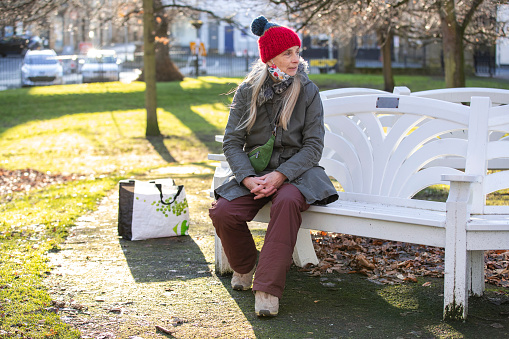 A mature caucasian woman wearing warm casual clothing. She is sitting on a park bench on a sunny winters day.