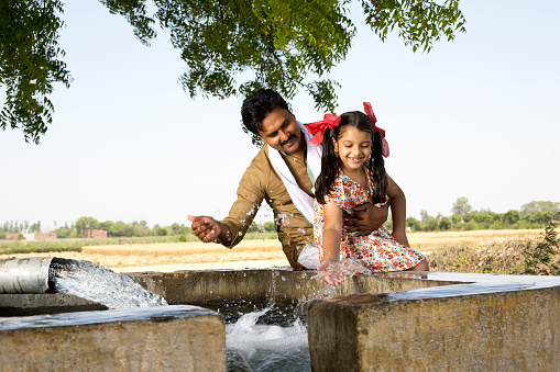 Rural Indian father and daughter having fun splashing water by pipe on agricultural field