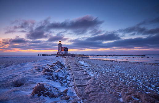 Paard van Marken photographed during sunset on a very cold winterday. The ground is covered in snow and the water is frozen. The sky is filled with clouds and the setting sun shines a bit through them.