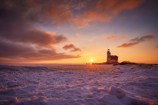 Paard van Marken photographed during sunset on a cold winterday. Everything is covered in snow and the water is frozen. The setting sun causes the sky to be beautifully colored. There are some clouds in the sky.
