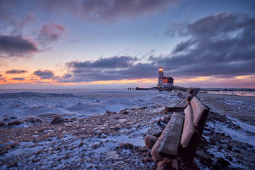 Paard van Marken photographed during sunset on a very cold winterday. The ground is covered in snow and the water is frozen. The sky is filled with clouds and the setting sun shines a bit through them. In the foreground are two benches that also have a layer of snow on them. The light of the lighthouse is on.