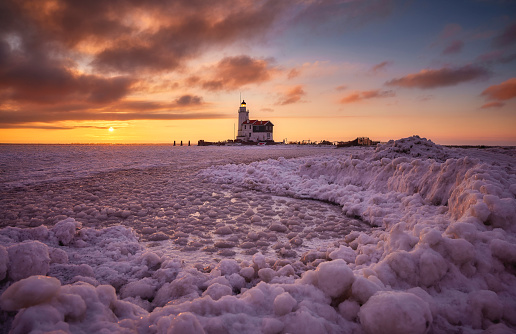 Paard van Marken photographed during sunset on a cold winterday. Everything is covered in snow and the water is totally frozen. The ice in the foreground is unusually shaped. The setting sun causes the sky to be beautifully colored. There are some clouds in the sky.