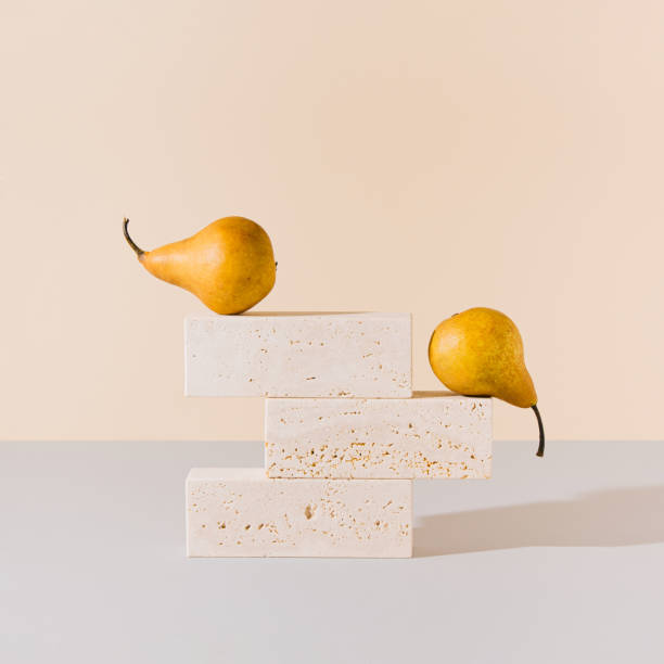 Yellow pears balancing on travertine marble blocks structure against gray and beige background. Creative summer fruit arrangement. Minimal retro styled surreal food art concept.