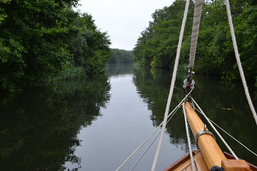 Peaceful, treelined river: green woodland reflected in tranquil water. The bowsprit and furled sail (jib) of the bow of a wooden yacht making its way up the centre; Norfolk Broads, Norfolk, UK