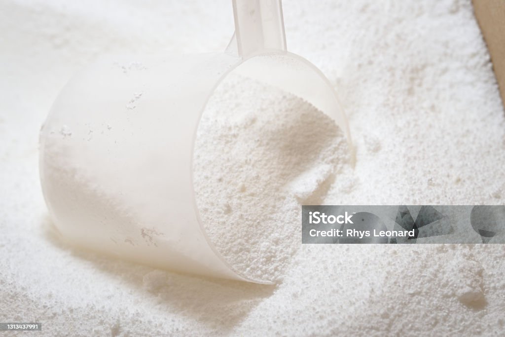 A scoop measures out laundry detergent powder for washing machine use. Close up of clear plastic scoop being used to measure white laundry detergent powder in a cardboard box. Bright image with macro powder detail Ground - Culinary Stock Photo