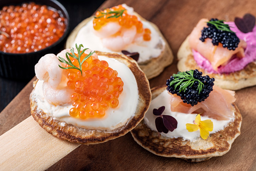 Selection of blinis made with; flour, buckwheat flour, egg yolks, yeast, milk and egg whites. Topped with prawns and sea trout roe, smoked salmon with beetroot infused creme fraiche and  black caviar and plain creme fraiche topped with salmon and caviar and decorated with wood sorrel and dill. Colour, horizontal format with some copy space.