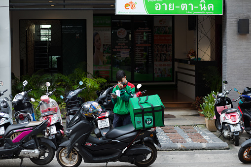Thai food delivery person arrived at a condominium in Bangkok ladprao. Young man is standing at motorcycle and is using mobile. Around several motorcycles are parked. Man is driving for 1112 express. He is wearing a face mask