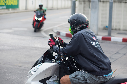 Thai man is waiting on motorcycle at junction and is using mobile. Man is wearing a helmet. In background a food express delivery person is crossing junction. Scene is in ladprao