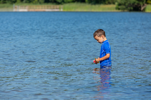 Side view of a young boy wading into the lake on a beautiful summer day at the beach. He is holding a small plastic sailboat toy in his hands.