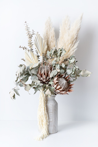 Beautiful dried flower arrangement in a stylish ceramic white vase. In the flower bunch is pink King Proteas, Palm Frond, Eucalyptus leaves, Amaranthus and Pampas grass, photographed on a white background.