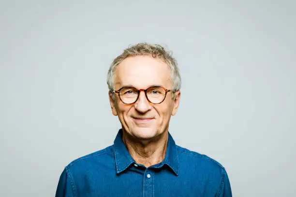 Portrait of elderly man wearing denim shirt and glasses smiling at camera. Confident senior entrepreneur, studio shot against grey background.