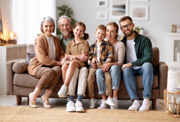 piękna wielopokoleniowa rodzina siedząca razem na kanapie w domu i uśmiechnięta przed kamerą - multi family zdjęcia i obrazy z banku zdjęć