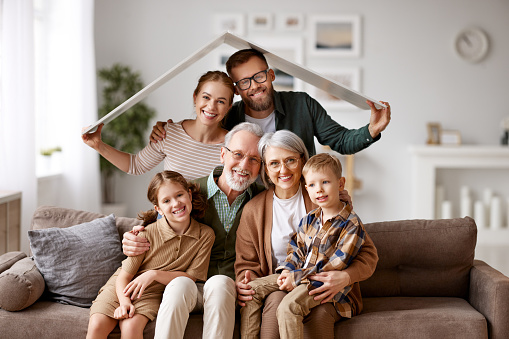 Big happy family, grandparents, mother, father with little kids son and daughter celebrating relocation in new home, sitting on the coach under paper roof and smiling at camera. Mortgage loan concept