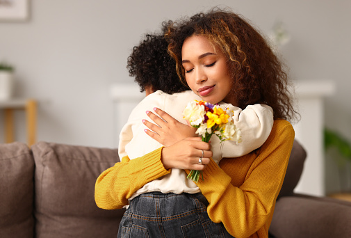 Little son congratulating mom with Mothers day at home and giving her fresh flower bouquet, happy mixed race woman mother embracing son while sitting on sofa. Family holidays and celebration concept