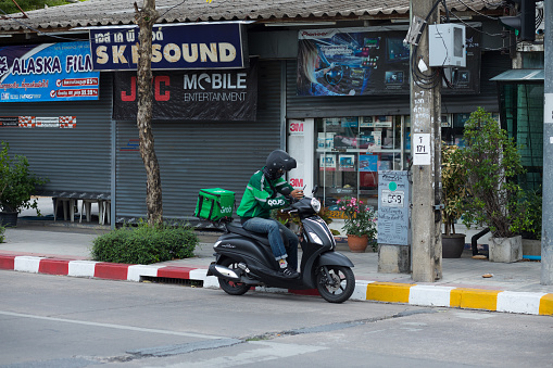 Thai man and delivery person is waiting with mobile on motorcycle in street Wanghin Road in Bangkok Ladprao. In background are several closed shops.