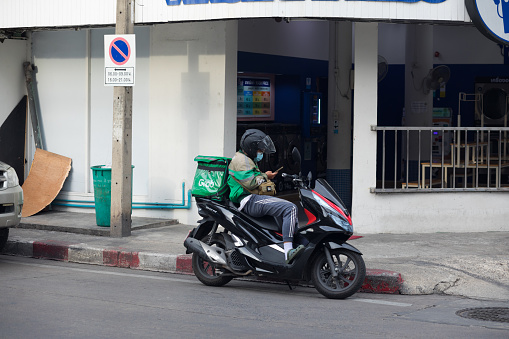 Thai express delivery person is sitting with mobile on motorcycle in street Chockchai 4 in Ladprao. In background is a laundry salon. Man is driving for Grab Food