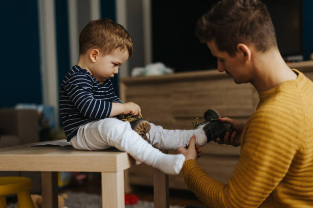 Putting on shoes Affectionate young father putting on shoes to his little son while he is sitting on the table in living room at home child tied up stock pictures, royalty-free photos & images