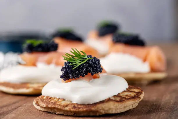 Homemade blinis made with; flour, buckwheat flour, egg yolks, yeast, milk and egg whites. Traditionally eaten with blinis you can create your own recipe, here shown with; creme fraiche, smoked salmon, caviar of course, topped with dill. Colour, horizontal format with some copy space.