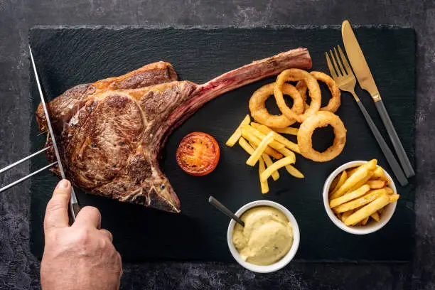 Overhead view of a huge, cooked tomahawk steak being served with; french fries, tomato slice, deep fried onion rings and béarnaise sauce as it is sliced at the table. Colour, horizontal with some copy space. The Tomahawk Steak got it’s name from their shape resembling a tomahawk axe. They have become popular in restaurants and with home barbecue chefs to see how much one person can eat!