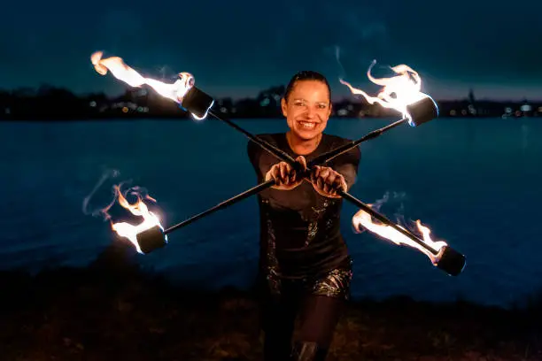 Female fire artist holding two flaming torches crossed at the camera. She ist looking at the camera and smiling.
