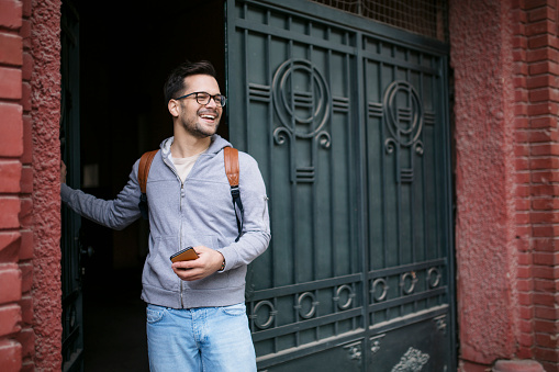Handsome young Caucasian man going outside for a walk, waring a backpack, smiling and using a smart phone