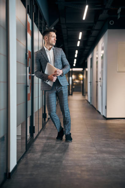 istock Stylish young man with laptop standing in office corridor 1313489139