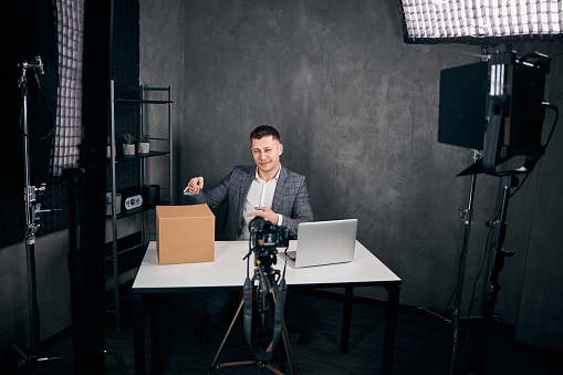 Smiling gentleman sitting at the table with laptop and pointing at cardboard box while filming video for blog