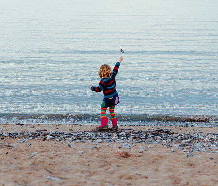 Color image depicting the rear view of a little girl, five years old, dressed in multi colored clothing. She is enthusiastically throwing a rock into the sea from the shoreline.