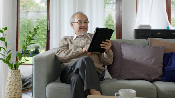 candido del vecchio pensionato asiatico che usa il tablet computer per trasferire denaro online o pagamento finanziario e leggere testo, sorridere o ridere con felice e positivo. asiatico senior con concetto di tecnologia digitale - ipad senior adult facebook sofa foto e immagini stock