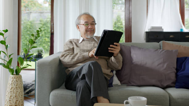candid of old asian retired man using tablet computer to transfer money online or financial payment and read text, smile or laugh with happy and positive. senior asian with digital technology concept - ipad senior adult facebook sofa imagens e fotografias de stock