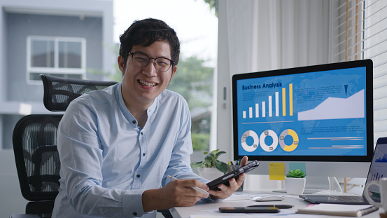 Retrato de la foto de cabeza de un joven asiático atractivo sentado sonriente trabajo ordenador de pantalla múltiple y tableta inteligente en el escritorio de la mesa en casa en el analista de datos independiente concepto, científico de datos para los n photo