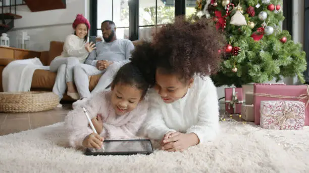X'mas Happy african family cute daughter, young sister or sibling having fun with smart tablet computer lying at home, little children using digital gadget learn to draw making sending e-card.