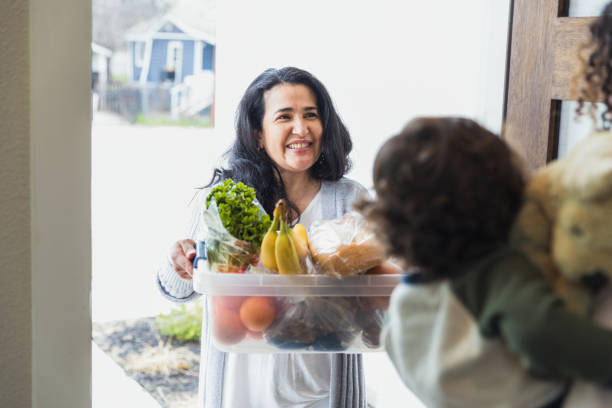 Friendly neighbor brings food to unrecognizable mother and child The friendly mature adult female neighbor smiles as she brings a box of fresh food to her unrecognizable neighbor. neighbour stock pictures, royalty-free photos & images