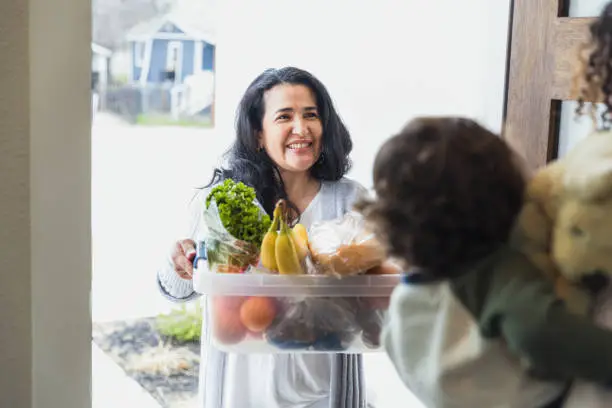 The friendly mature adult female neighbor smiles as she brings a box of fresh food to her unrecognizable neighbor.