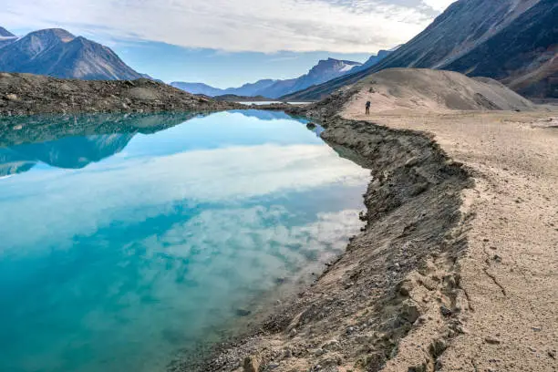 Photo of Hiker walking by a lake through remote arctic valley on a partly cloudy summer day. Dramatic arctic landscape of Akshayuk Pass, Baffin Island, Canada. Enormous glacier moraine.