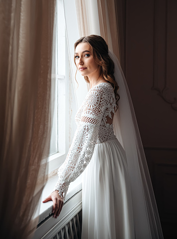 A young bride in a beautiful boho lace dress stands at the window. Wedding photo session, bride's morning.