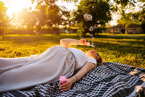 Young pregnant woman lying down in public park and blowing soap bubbles.