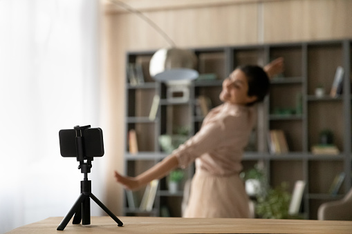Close up focus of modern smartphone camera shoot ethnic woman dancing in background recording video blog or live broadcast. Cellphone gadget on tripod catch female dancing. Technology concept.
