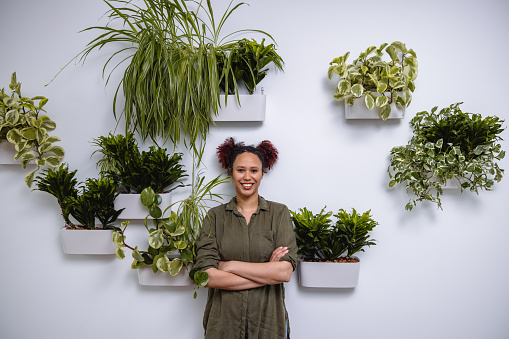 Portrait of a beautiful mixed race businesswoman in 20's smiling with hands crossed. Standing in a modern office in front of a plant wall.