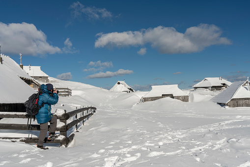 Men photographing idyllic landscape in Velika planina, Big Pasture Plateau in Slovenia, Europe, wooden pasture cottage covered with lot of snow, mountains against clear blue sky.