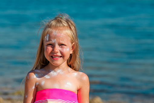 A beautiful little girl smiles in a swimsuit standing by the sea with sunscreen smeared on her face. Copy space