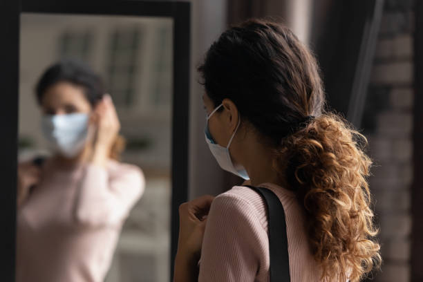 careful young female putting medical mask on face by mirror - woman in mirror backview imagens e fotografias de stock
