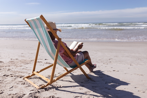 Senior african american woman reading a book while sitting on deck chair at the beach. travel vacation retirement lifestyle concept
