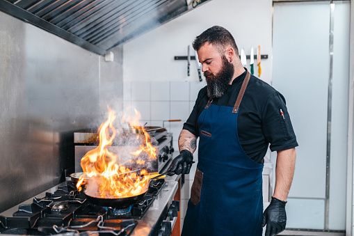Professional Chef fires up oil on a cooking pen. Bearded young chef cooks food with fire at kitchen restaurant.