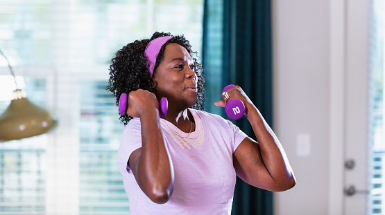 A senior African-American woman in her 60s working out at home in the living room. She is lifting handweights, taking an online exercise class, looking at a screen which is off-camera.