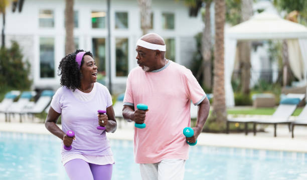 Senior African-American couple exercising on pool deck A senior African-American couple exercising together on a pool deck. They are staying fit and active, having fun during retirement. The woman, in her 60s, and her partner, in his 70s, are looking at each other as they power walk with hand weights. body positive couple stock pictures, royalty-free photos & images